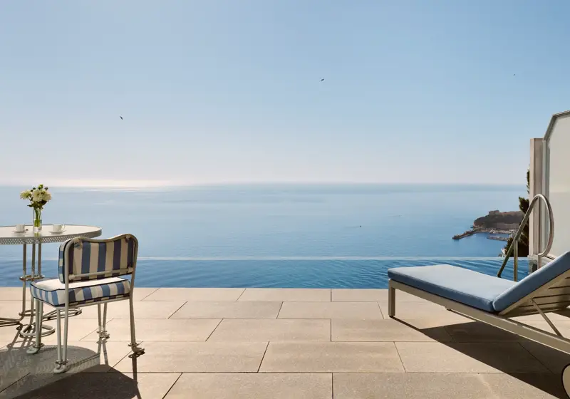 Terrasse avec chaise rayée, table et transat donnant sur une piscine à débordement et une vue panoramique sur la mer sous un ciel bleu clair.