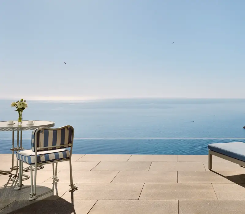 Terrace with striped chair, table, and sun lounger overlooking an infinity pool and panoramic sea view under a clear blue sky.