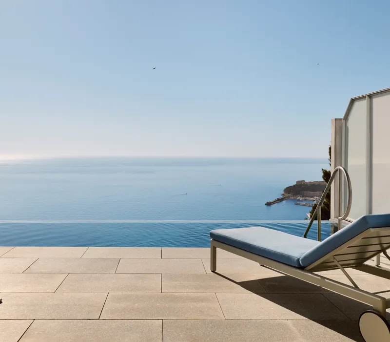 Terrasse avec chaise rayée, table et transat donnant sur une piscine à débordement et une vue panoramique sur la mer sous un ciel bleu clair.