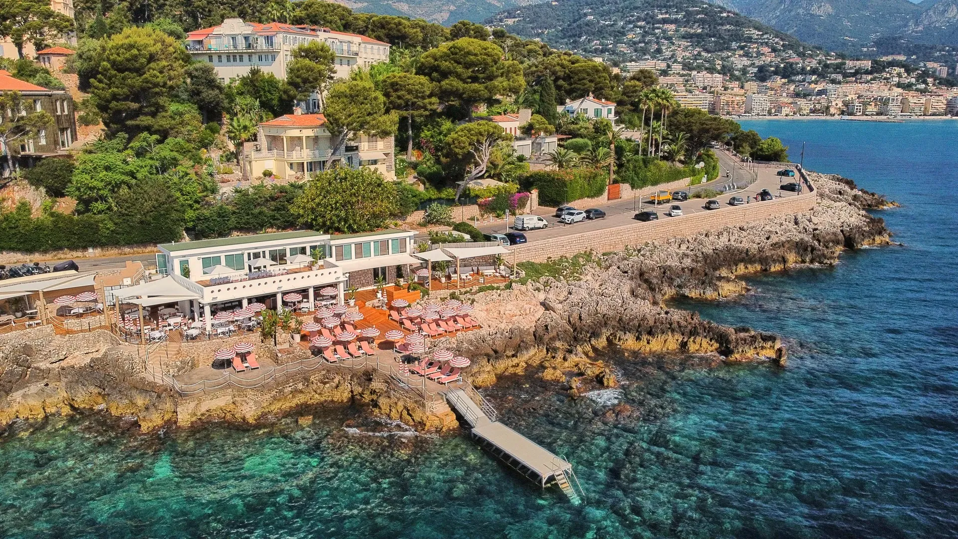 Restaurant en bord de mer avec parasols roses et terrasse sur rochers, entouré de verdure, dominant les eaux turquoise et la ville côtière.