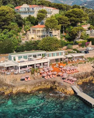 Restaurant en bord de mer avec parasols roses et terrasse sur rochers, entouré de verdure, dominant les eaux turquoise et la ville côtière.