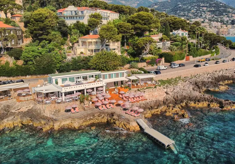 Seaside restaurant with pink parasols and terrace seating on rocky shoreline, surrounded by greenery, overlooking turquoise waters and coastal town.