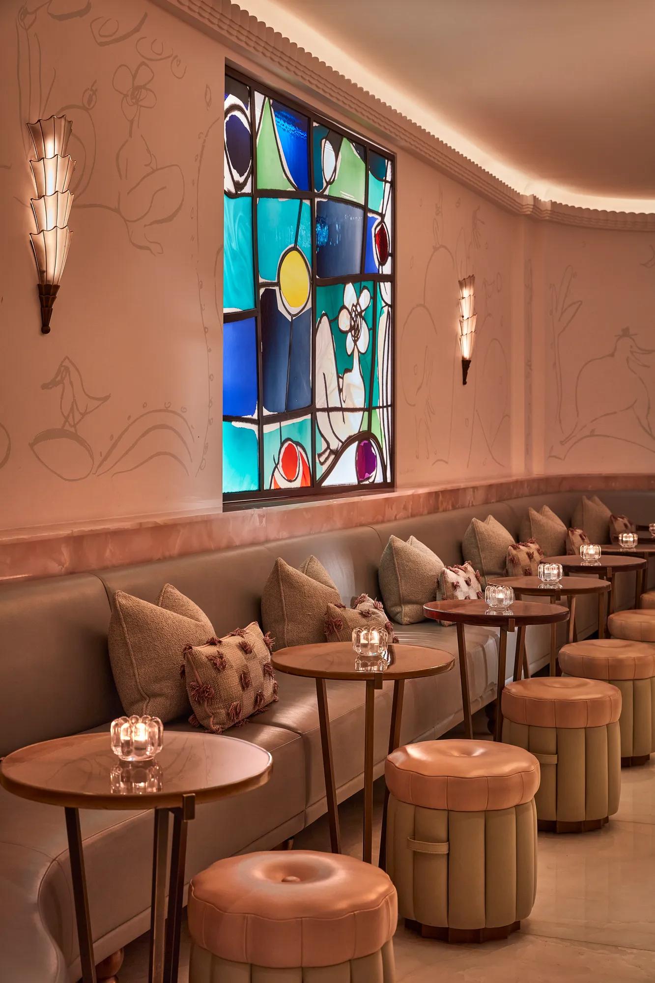 Elegant bar lounge with soft leather seating, circular tables, and art deco wall sconces. A colourful stained-glass window adds warmth and vibrancy to the pastel-toned space decorated with subtle wall engravings.