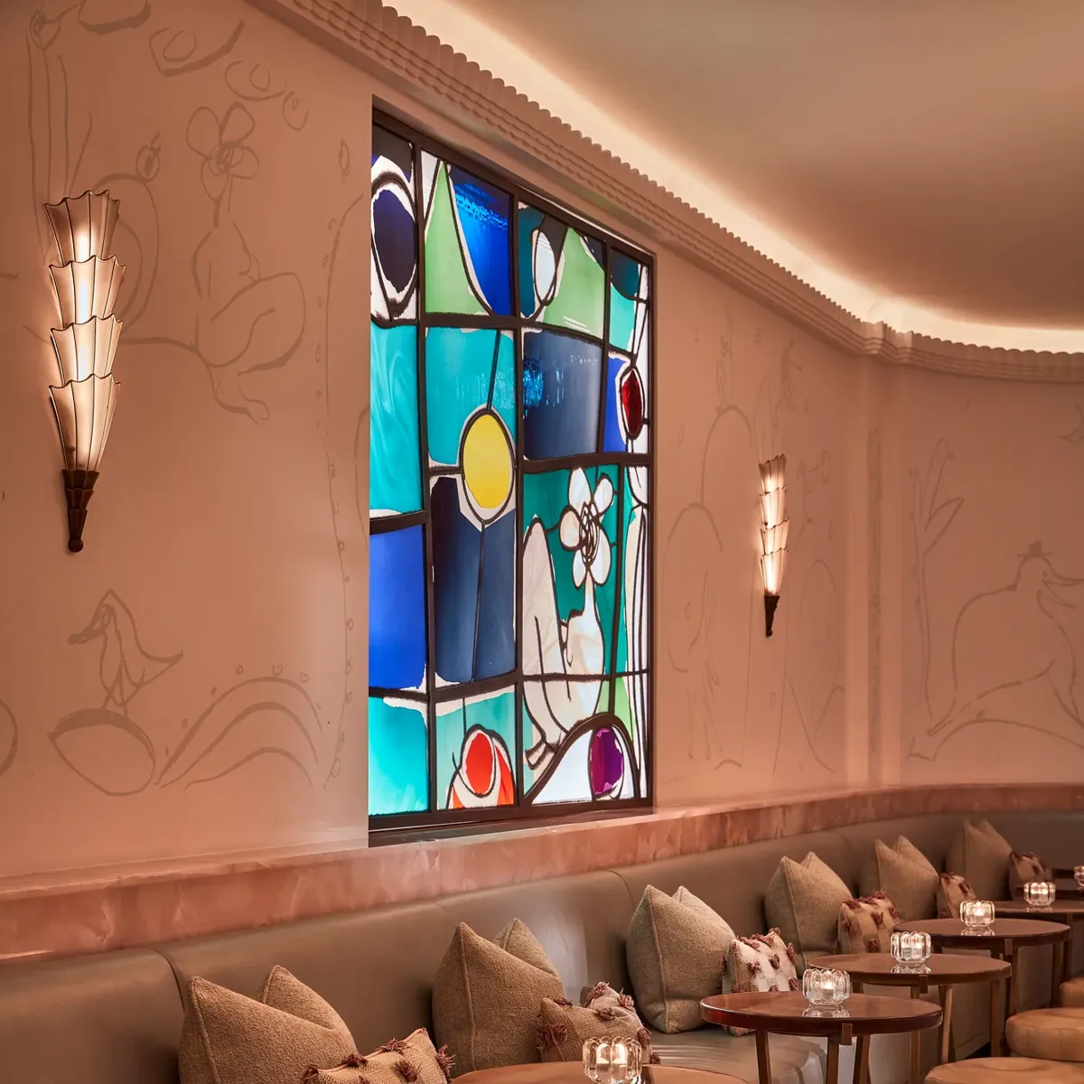 Elegant bar lounge with soft leather seating, circular tables, and art deco wall sconces. A colourful stained-glass window adds warmth and vibrancy to the pastel-toned space decorated with subtle wall engravings.