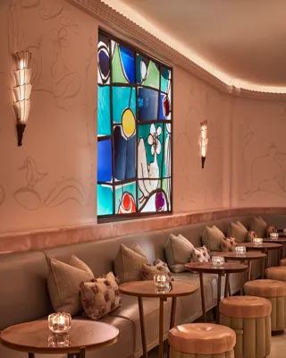 Elegant bar lounge with soft leather seating, circular tables, and art deco wall sconces. A colourful stained-glass window adds warmth and vibrancy to the pastel-toned space decorated with subtle wall engravings.