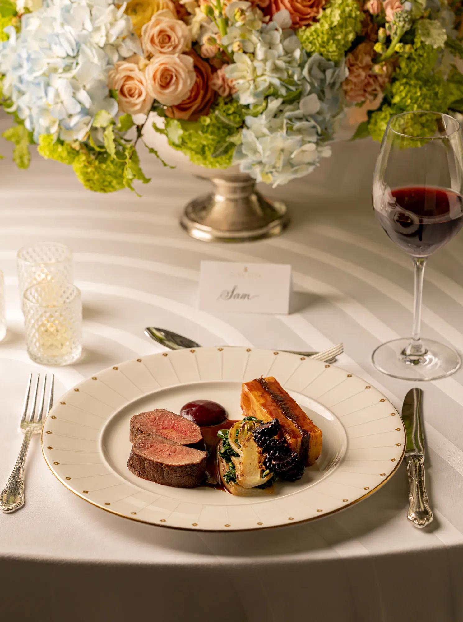 Sliced roasted lamb with glazed vegetables, beetroot purée and seasonal greens, served on fine china at an elegantly set dining table with floral centrepiece and red wine.