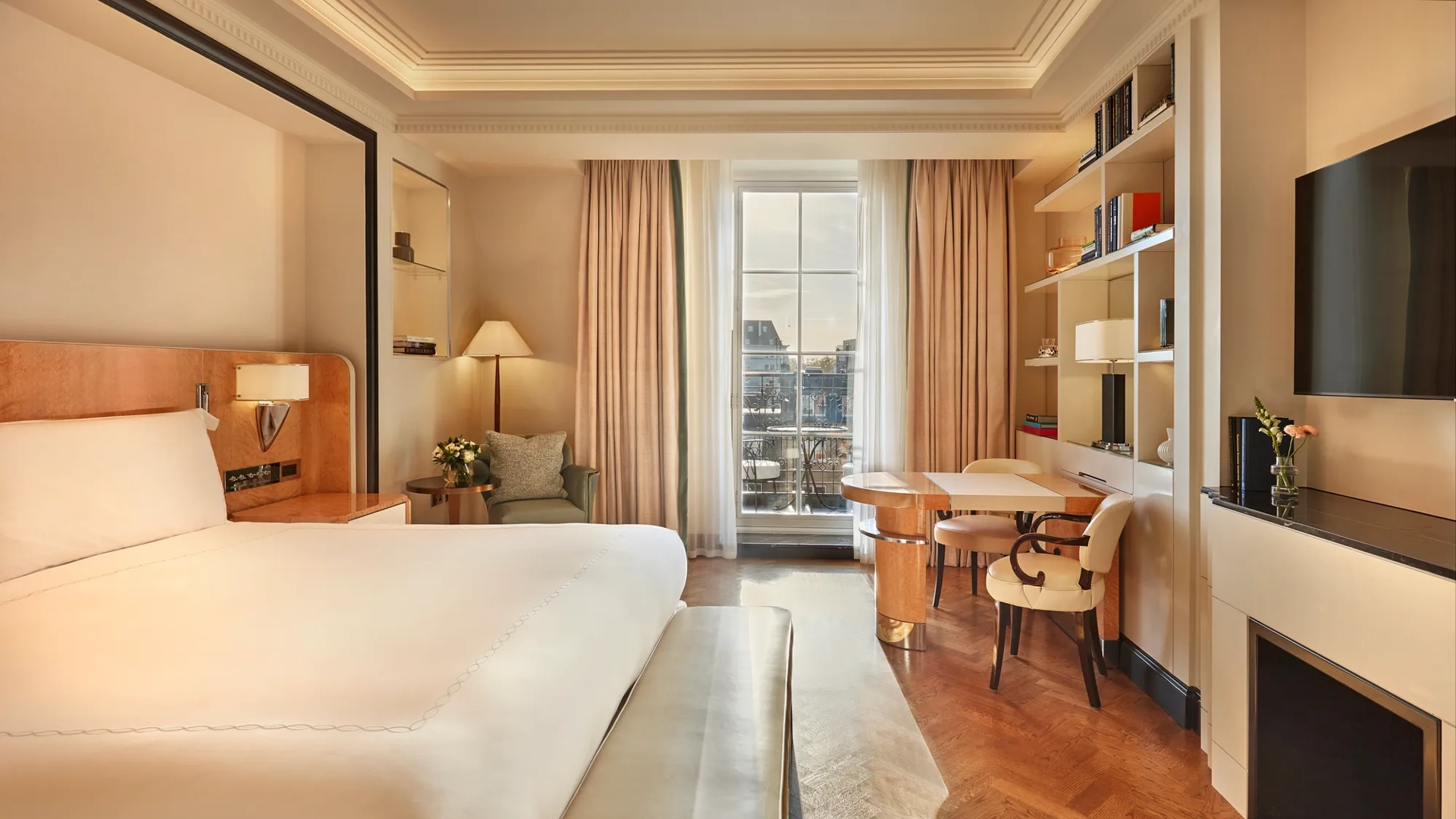 Elegant bedroom with a large bed, cream and wood accents, a writing desk with chairs by the window, and soft natural light illuminating a private balcony view.