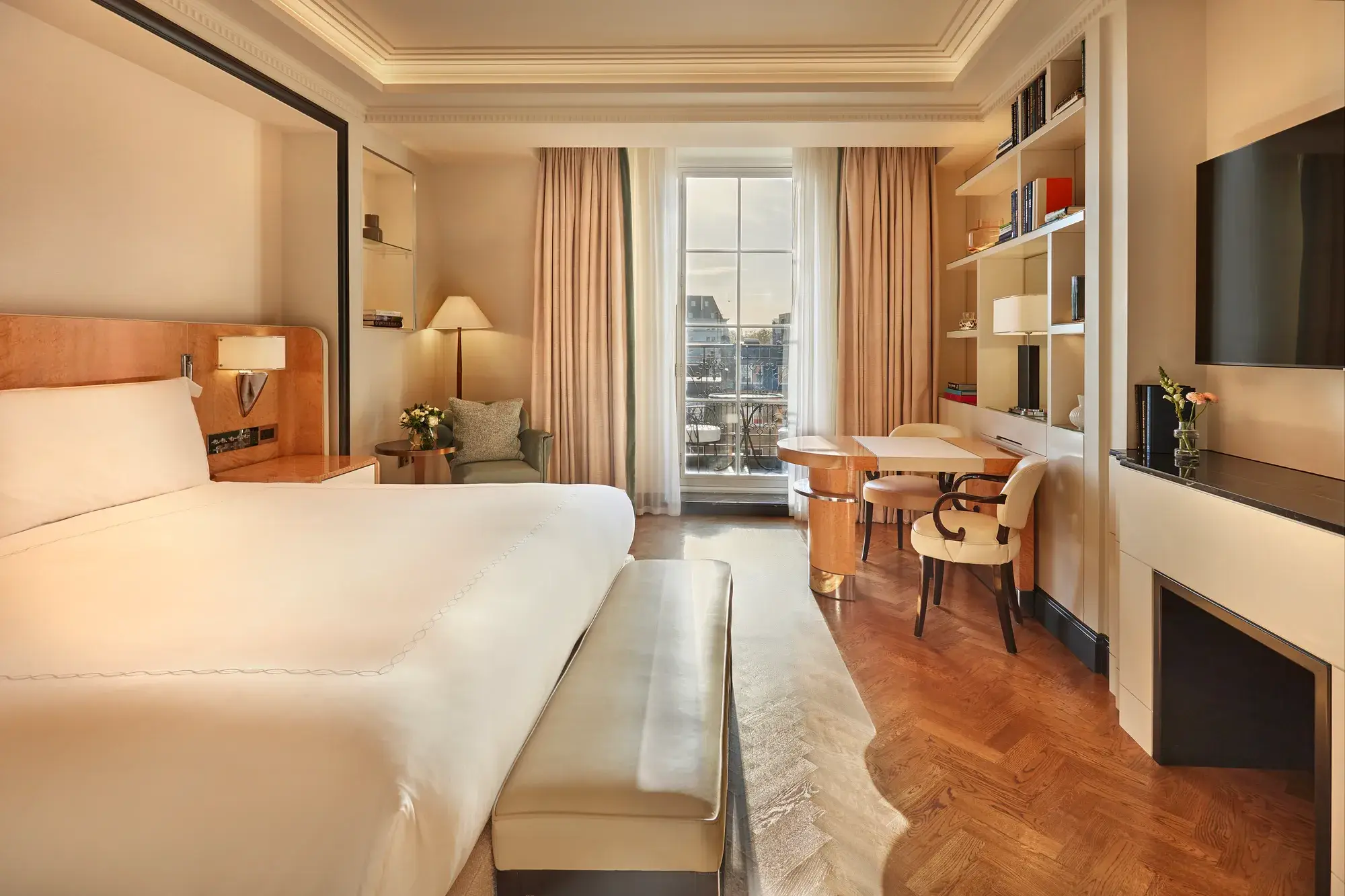 Elegant bedroom with a large bed, cream and wood accents, a writing desk with chairs by the window, and soft natural light illuminating a private balcony view.