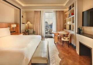 Elegant bedroom with a large bed, cream and wood accents, a writing desk with chairs by the window, and soft natural light illuminating a private balcony view.