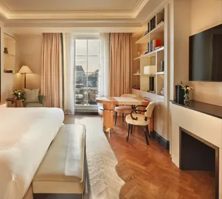 Elegant bedroom with a large bed, cream and wood accents, a writing desk with chairs by the window, and soft natural light illuminating a private balcony view.