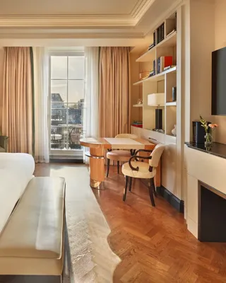 Elegant bedroom with a large bed, cream and wood accents, a writing desk with chairs by the window, and soft natural light illuminating a private balcony view.
