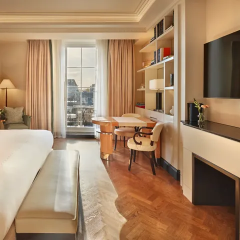 Elegant bedroom with a large bed, cream and wood accents, a writing desk with chairs by the window, and soft natural light illuminating a private balcony view.