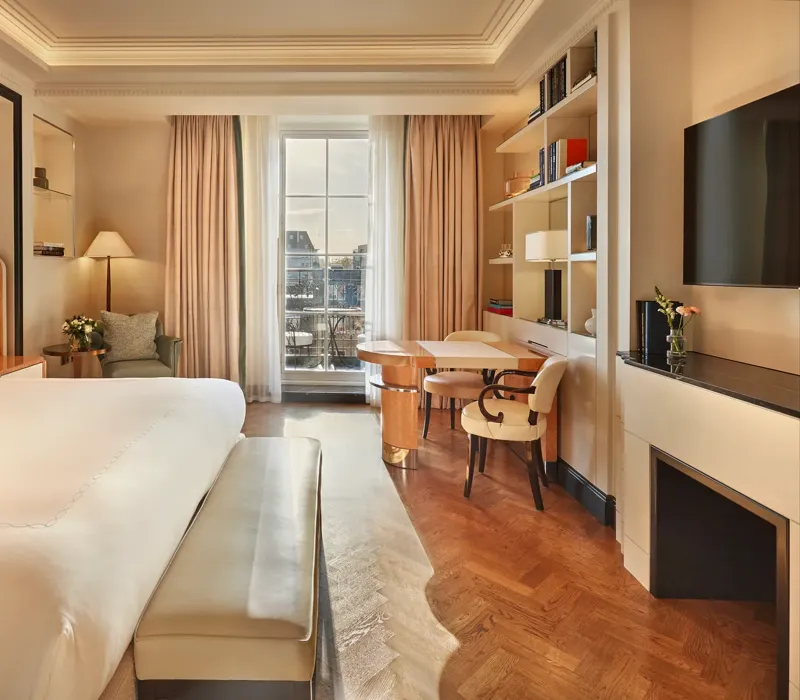 Elegant bedroom with a large bed, cream and wood accents, a writing desk with chairs by the window, and soft natural light illuminating a private balcony view.