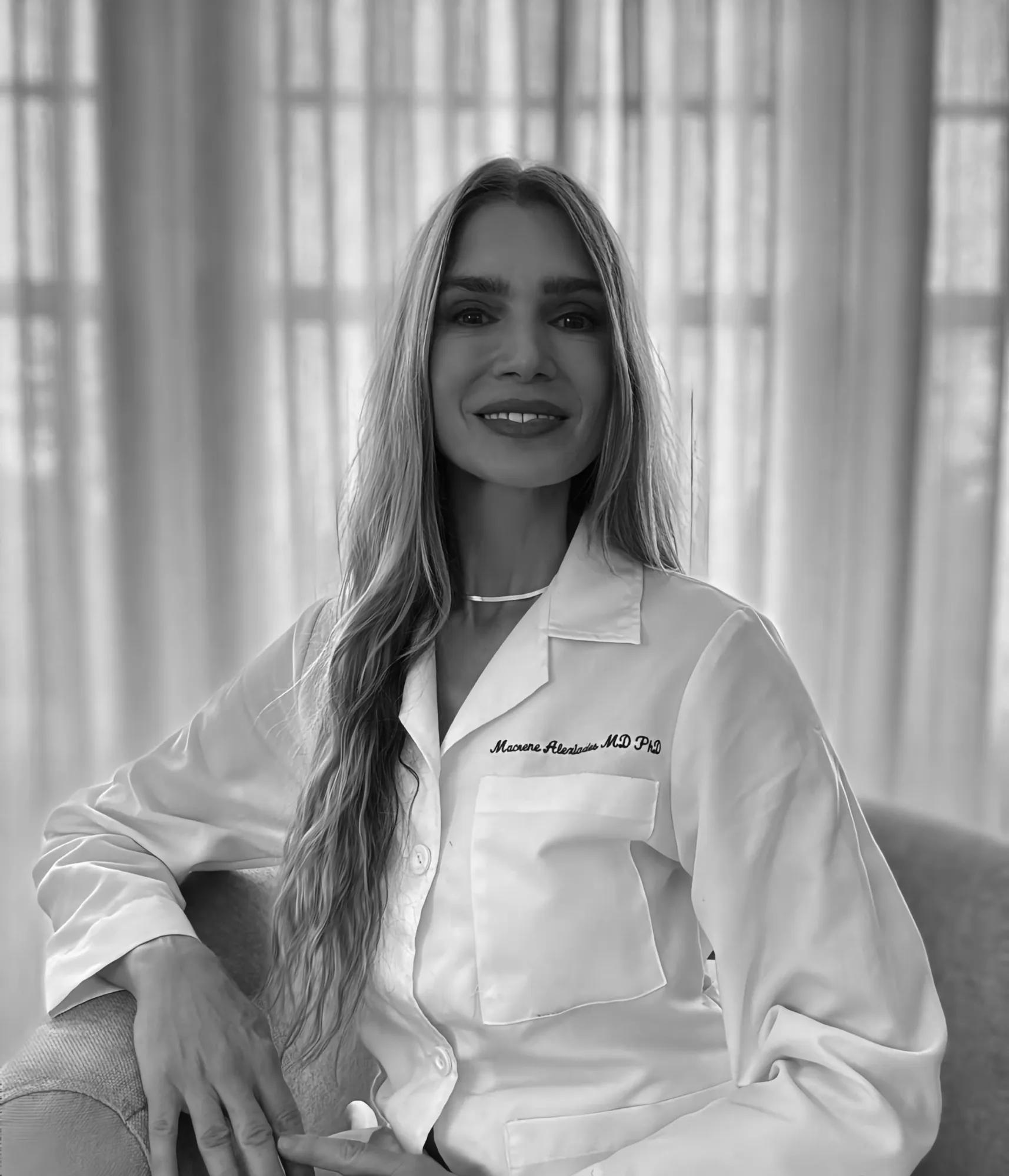 Portrait of a smiling woman in a white lab coat, seated in front of sheer curtains.