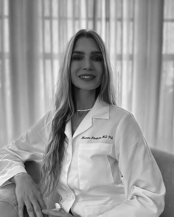 Portrait of a smiling woman in a white lab coat, seated in front of sheer curtains.
