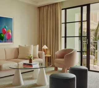 Bright living area with cream sofa, tub chair, glass coffee table, two ottomans, and balcony doors; framed balloons artwork on the wall.