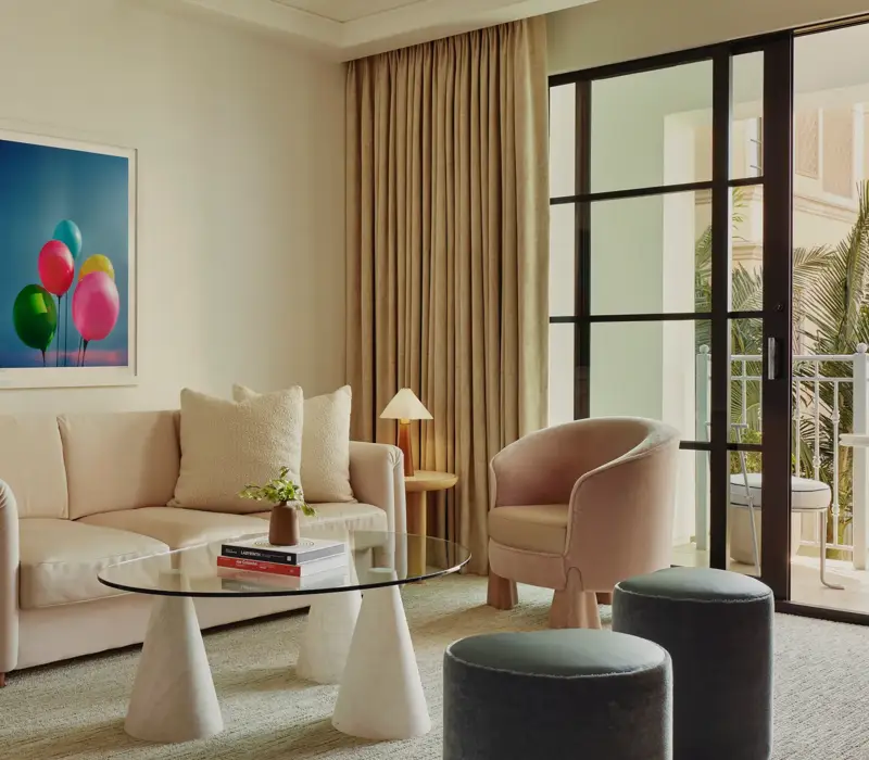 Bright living area with cream sofa, tub chair, glass coffee table, two ottomans, and balcony doors; framed balloons artwork on the wall.