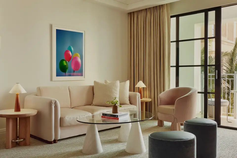 Bright living area with cream sofa, tub chair, glass coffee table, two ottomans, and balcony doors; framed balloons artwork on the wall.