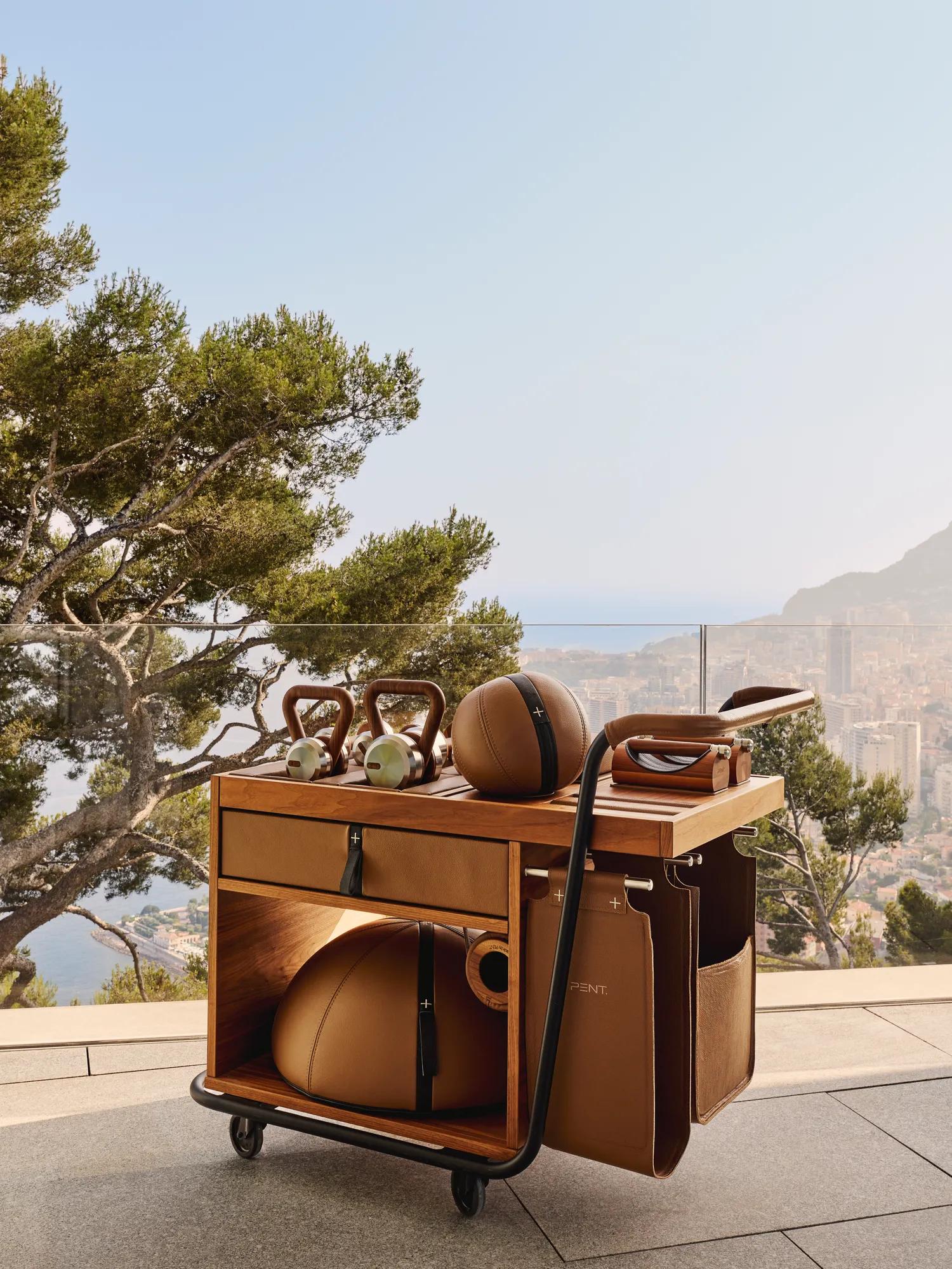 Wooden fitness trolley with weights and equipment on terrace, overlooking coastal landscape with trees, sea and distant hillside buildings in sunlight.