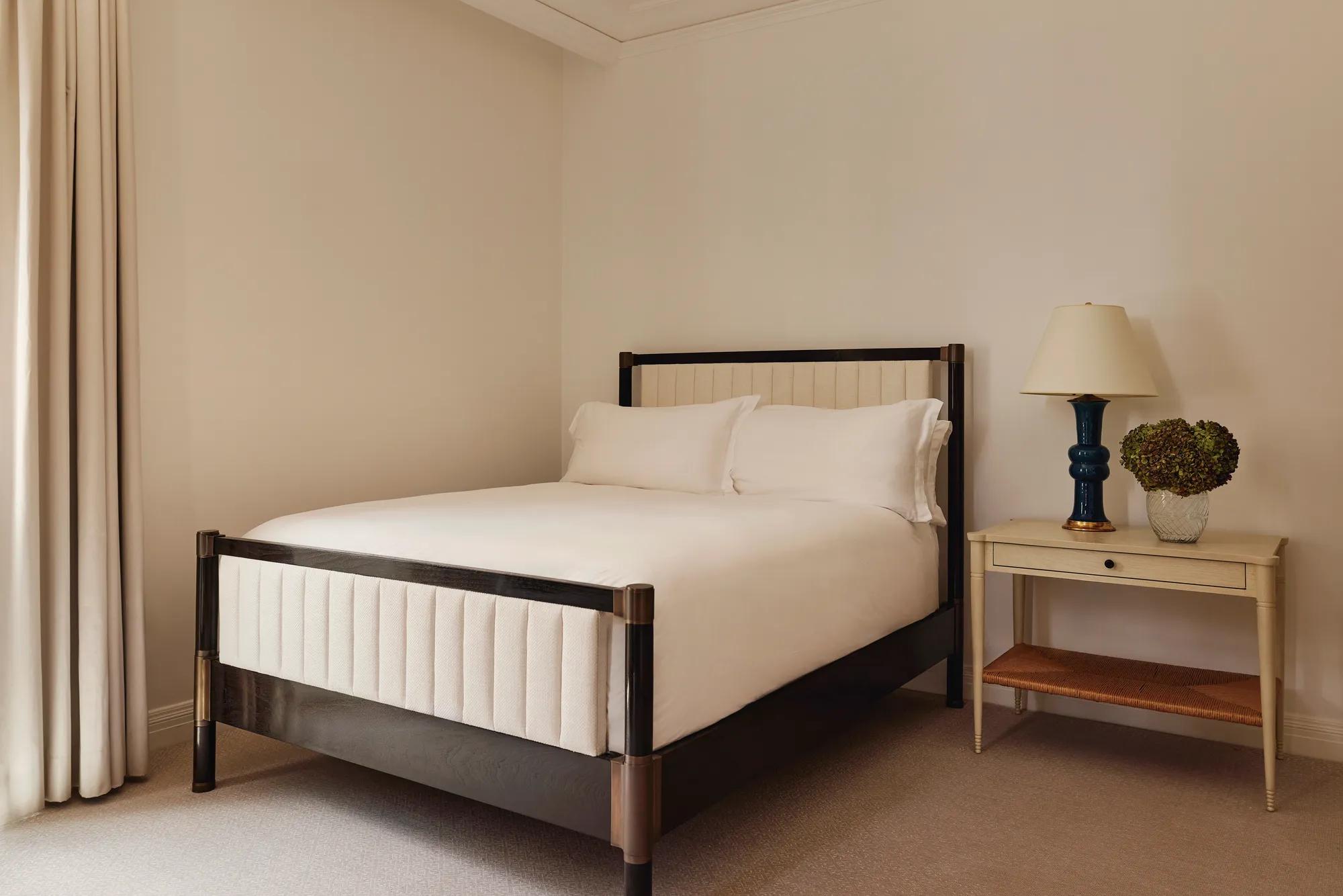 Minimalist bedroom with a cream-upholstered double bed, white bedding, and a side table with a navy ceramic lamp and glass vase of hydrangeas.