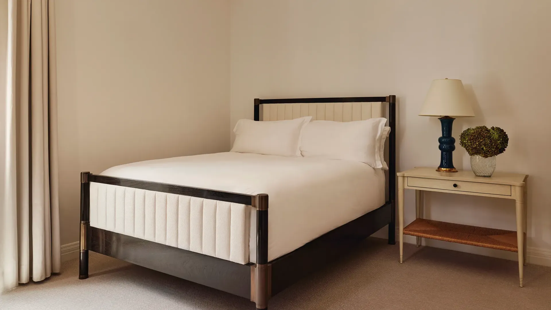Minimalist bedroom with a cream-upholstered double bed, white bedding, and a side table with a navy ceramic lamp and glass vase of hydrangeas.