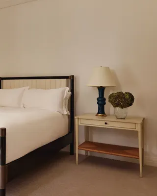 Minimalist bedroom with a cream-upholstered double bed, white bedding, and a side table with a navy ceramic lamp and glass vase of hydrangeas.