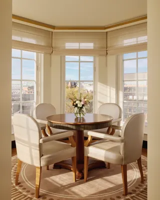 Bright corner dining area with four cream upholstered chairs around a round wooden table, a vase of flowers in the centre, and large windows offering city views.