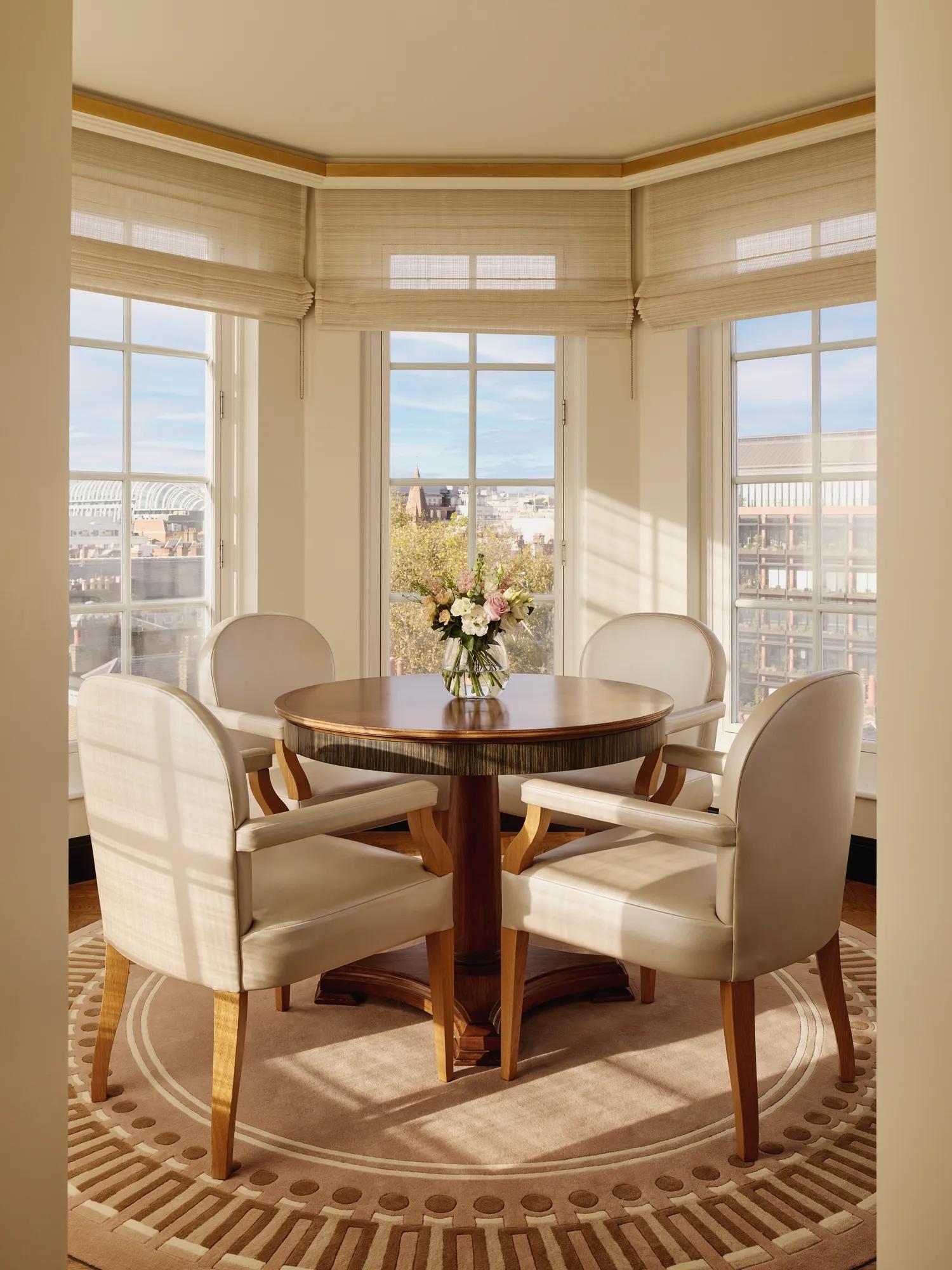 Bright corner dining area with four cream upholstered chairs around a round wooden table, a vase of flowers in the centre, and large windows offering city views.