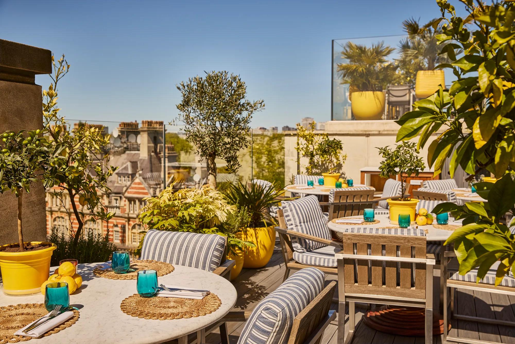Sunlit rooftop terrace with striped seating, marble tables, potted plants and city views beyond glass railings.