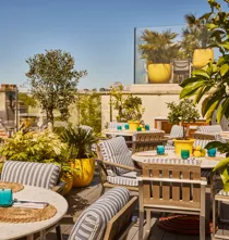 Sunlit rooftop terrace with striped seating, marble tables, potted plants and city views beyond glass railings.