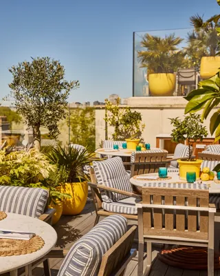 Sunlit rooftop terrace with striped seating, marble tables, potted plants and city views beyond glass railings.