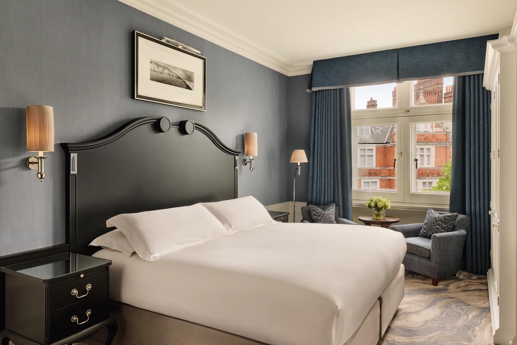 Bedroom with large bed and dark headboard, blue walls, and seating by a window overlooking red-brick rooftops.