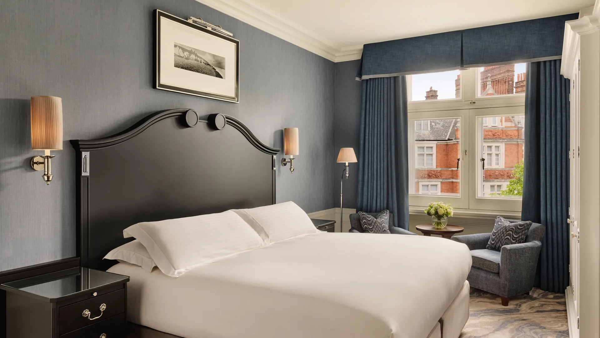 Bedroom with large bed and dark headboard, blue walls, and seating by a window overlooking red-brick rooftops.