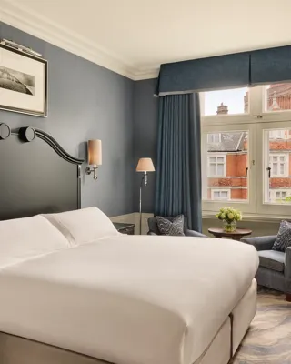 Bedroom with large bed and dark headboard, blue walls, and seating by a window overlooking red-brick rooftops.