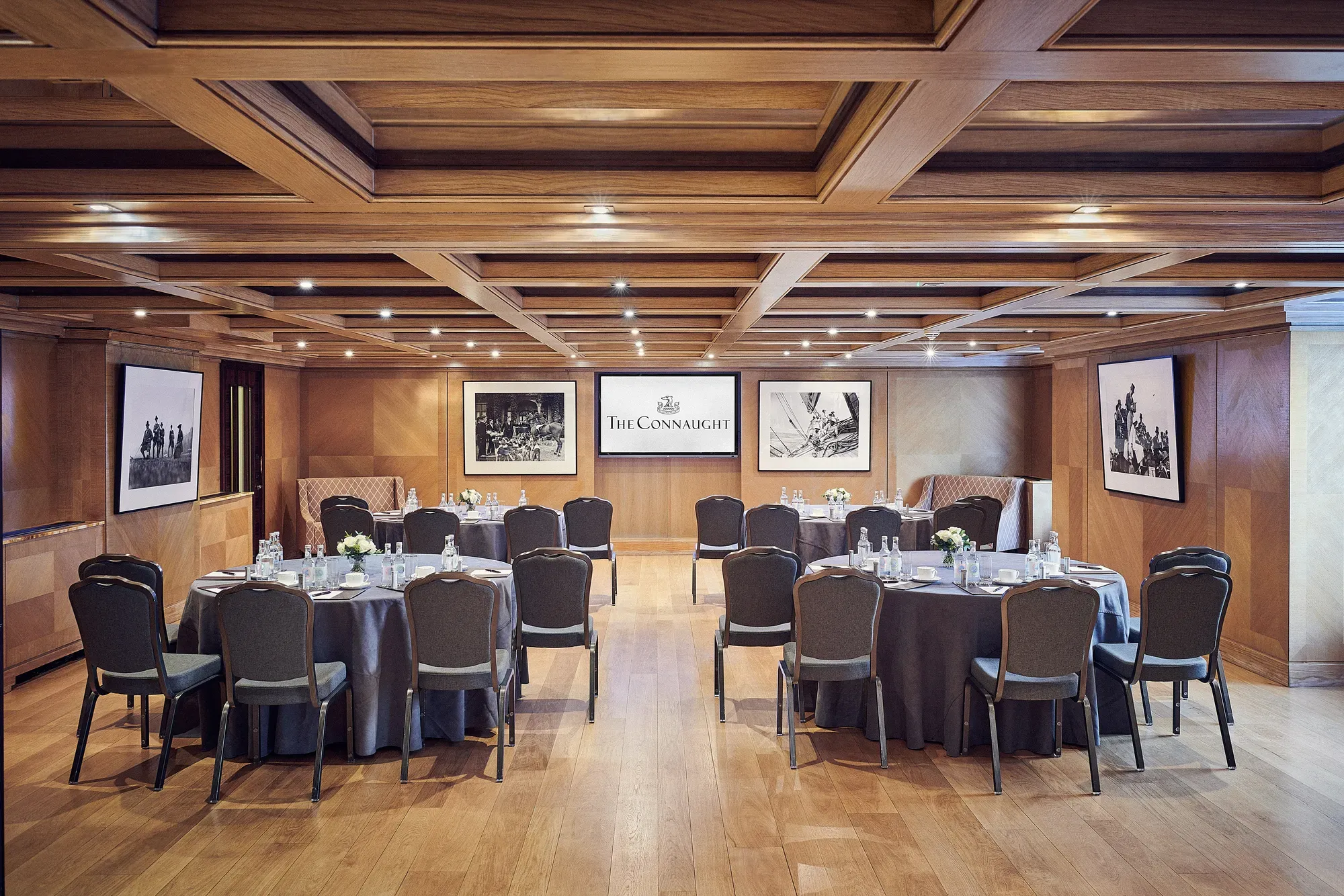 Wood-paneled meeting room with round banquet tables, black chairs, and Connaught presentation screen.