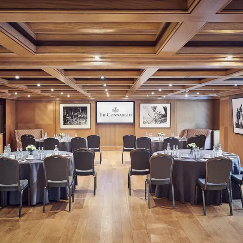 Wood-paneled meeting room with round banquet tables, black chairs, and Connaught presentation screen.