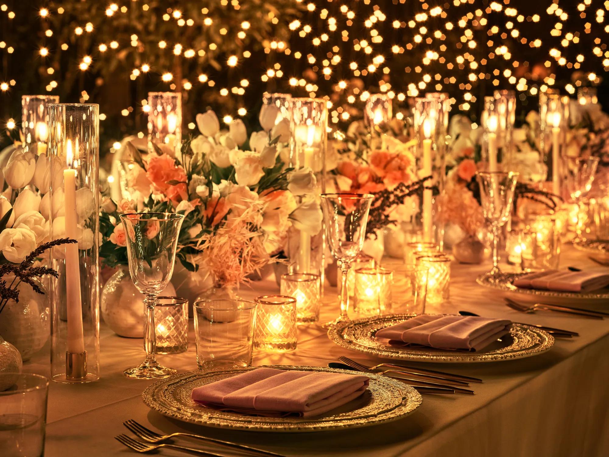 Warm, candlelit garden tablescape at night; gold glow on crystal glassware, flowers, and chargers with bokeh string lights behind.