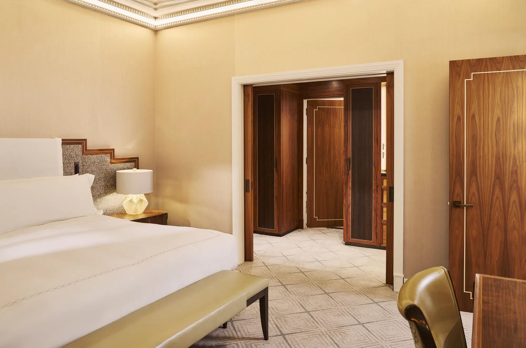 Elegant bedroom with white bed, bedside lamp, upholstered bench, and dark wood doors opening to marble hall.