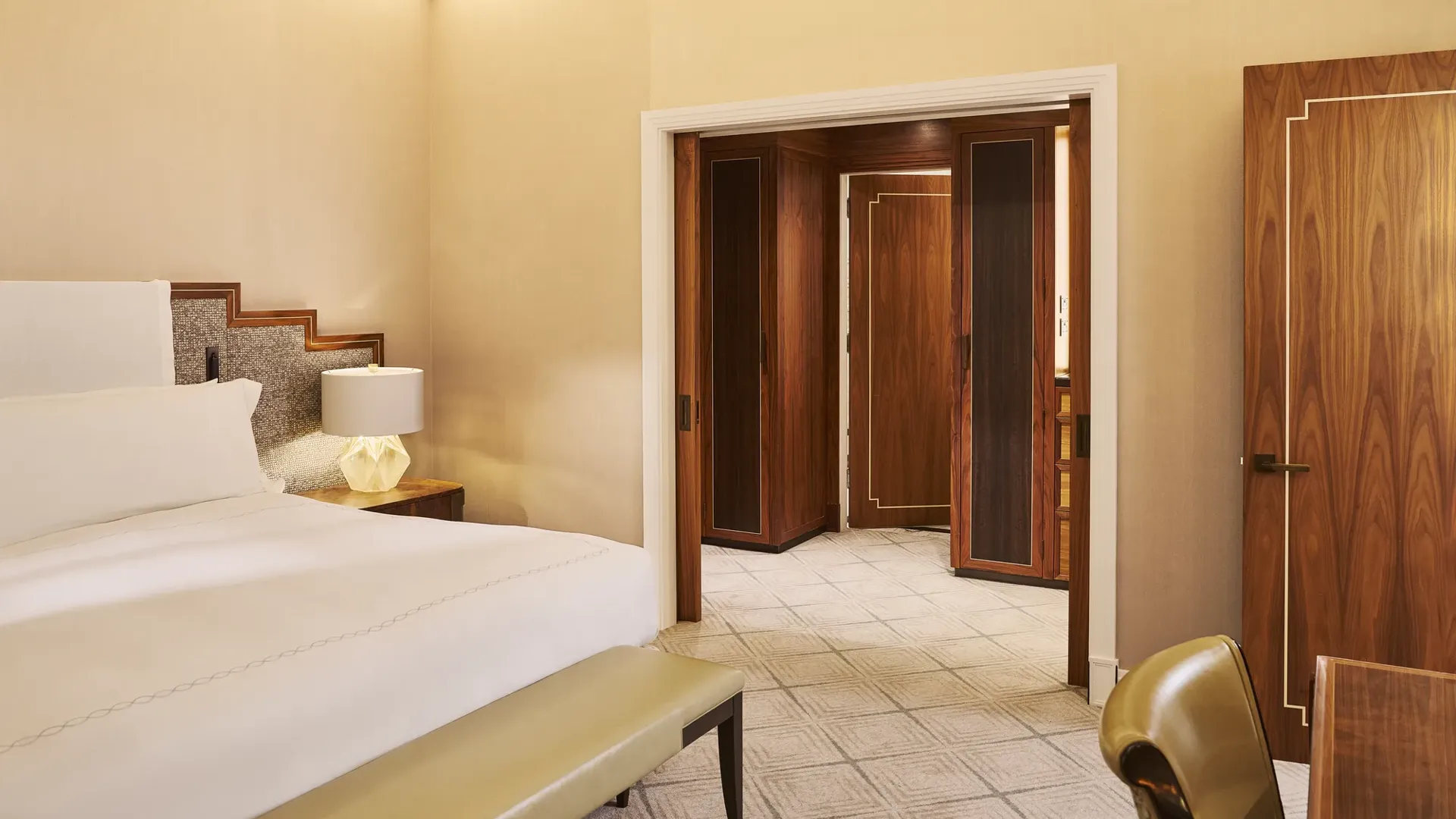 Elegant bedroom with white bed, bedside lamp, upholstered bench, and dark wood doors opening to marble hall.