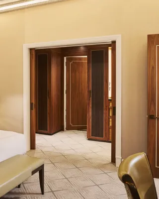 Elegant bedroom with white bed, bedside lamp, upholstered bench, and dark wood doors opening to marble hall.