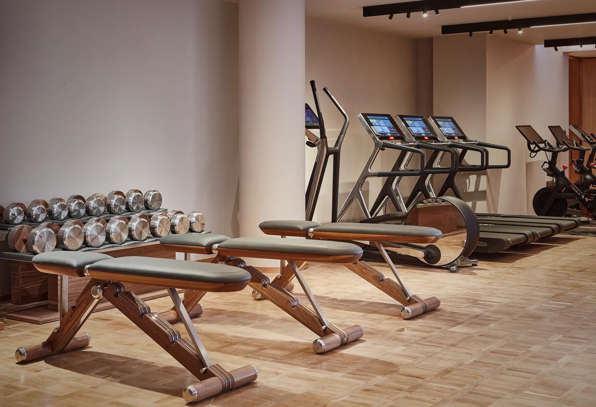 Modern fitness studio at The Maybourne Riviera featuring wooden exercise benches, polished dumbbells, treadmills, and stationary bikes on a parquet floor.