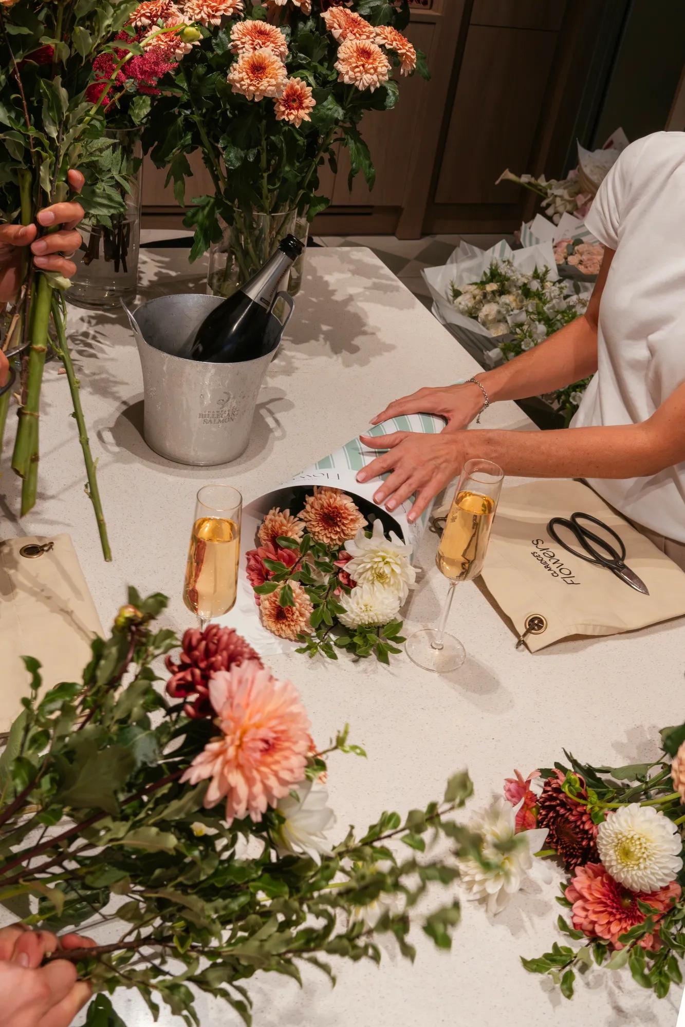Floristry workshop in progress at Claridge’s Flowers, with seasonal blooms, champagne and bouquet-making materials displayed on a worktable.