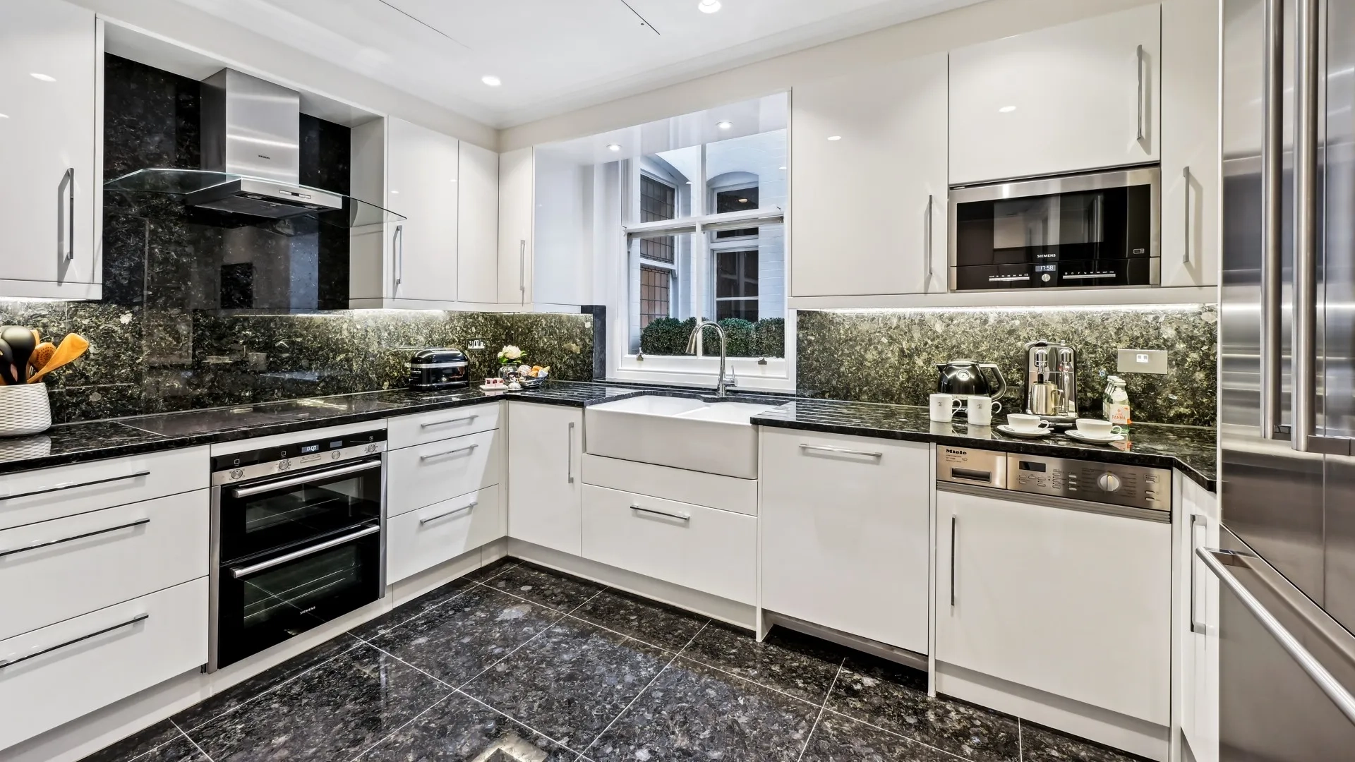 Modern white kitchen with black marble counters, built-in appliances, and a window over the sink.