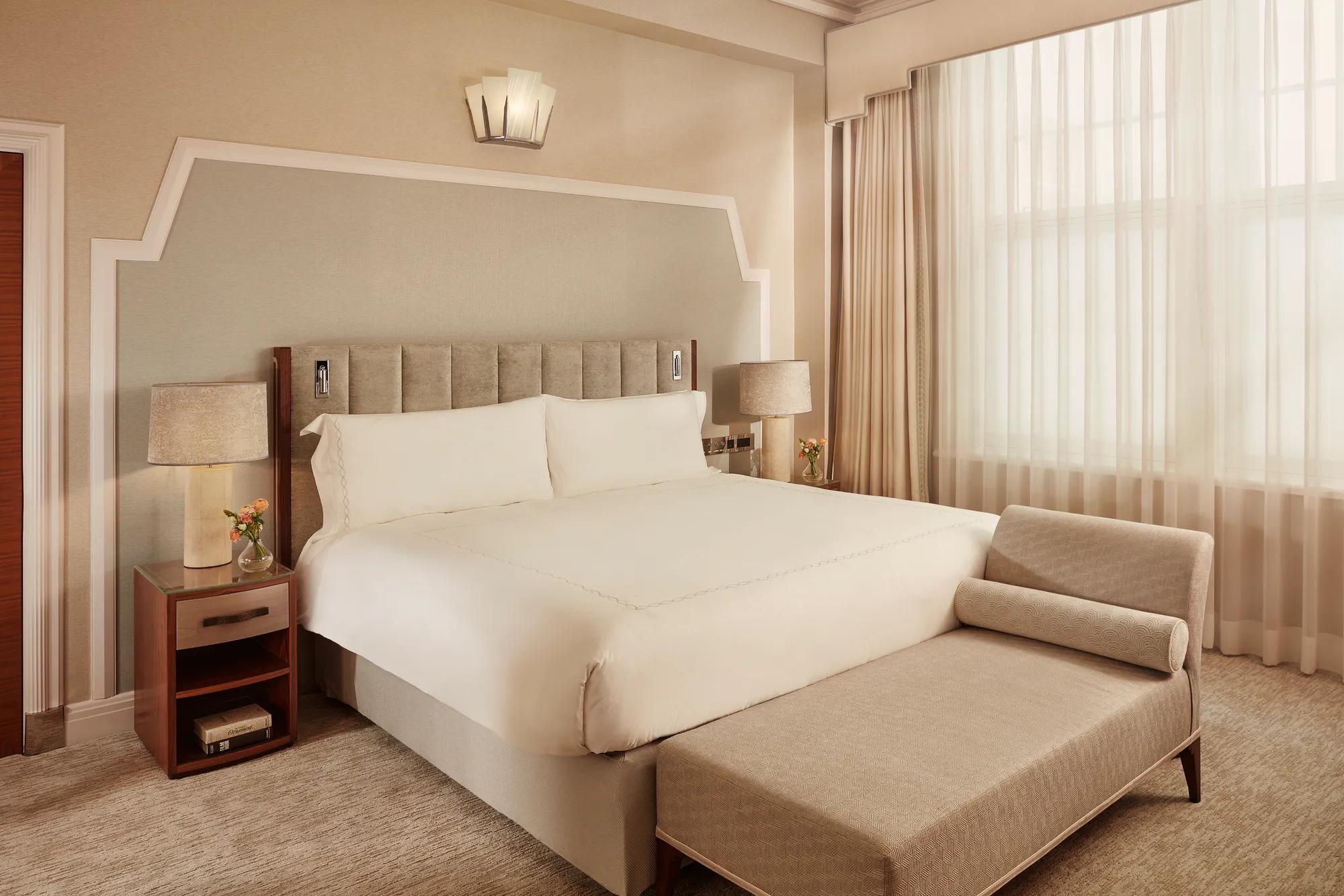 Elegant bedroom with king bed, bedside tables, chaise lounge, and large curtained window.