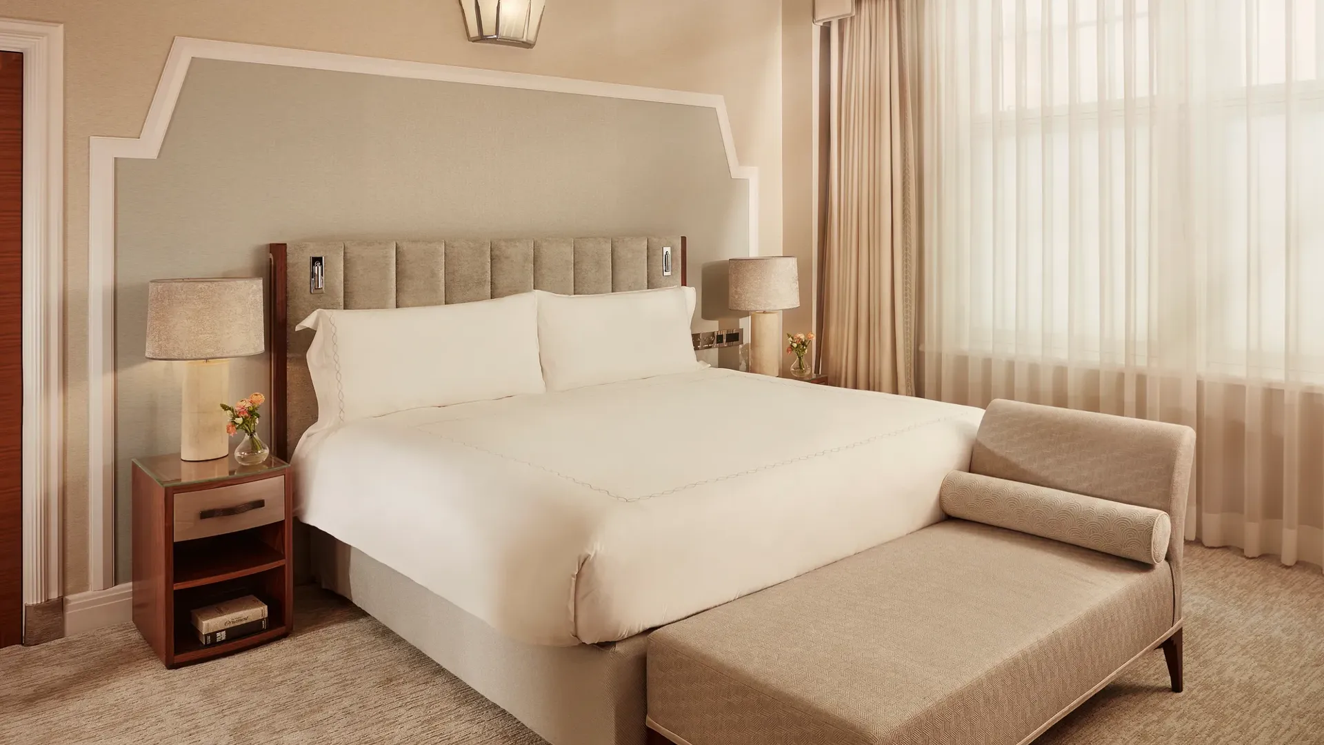 Elegant bedroom with king bed, bedside tables, chaise lounge, and large curtained window.