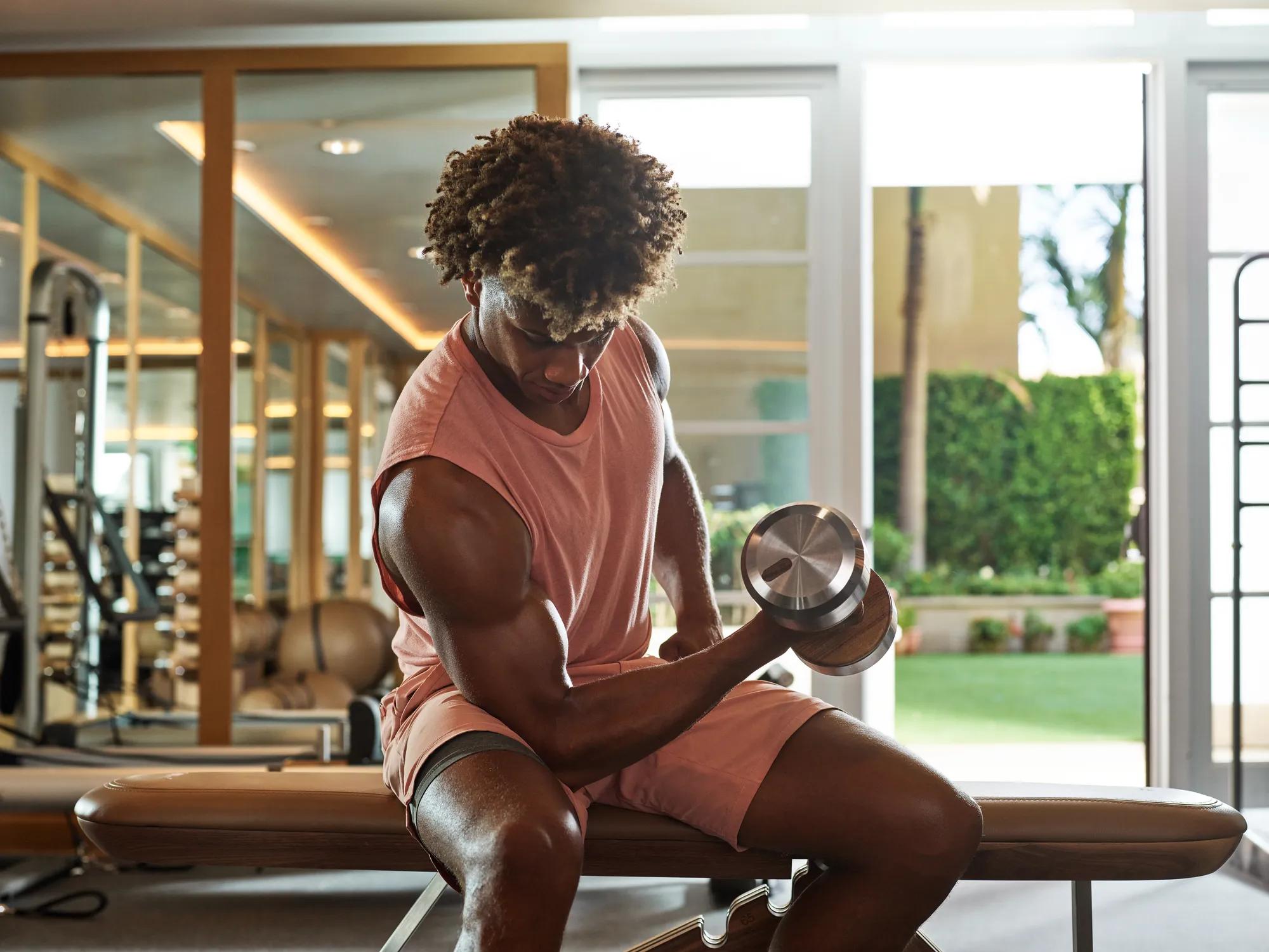 Man lifting dumbbell while seated on bench inside Maybourne Beverly Hills fitness center with mirrored walls