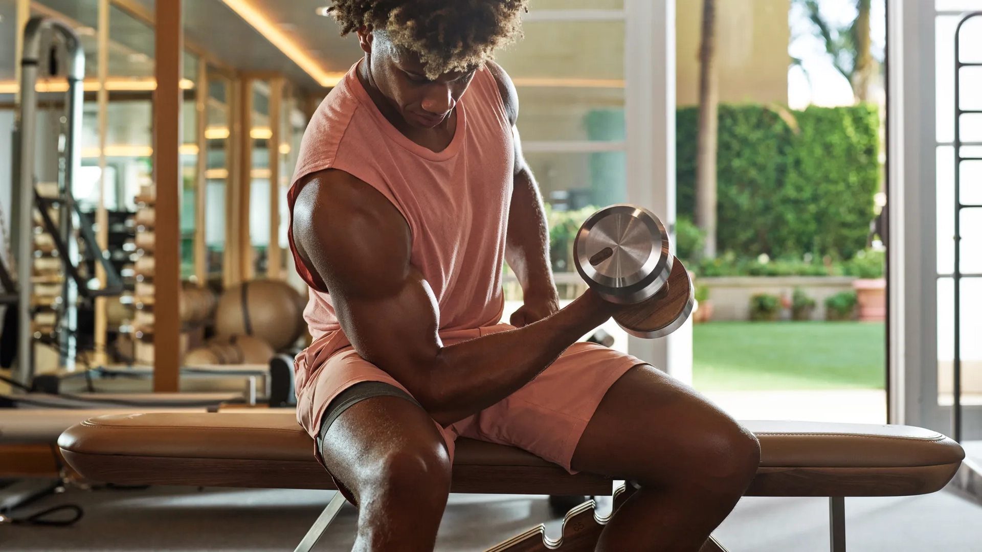 Man lifting dumbbell while seated on bench inside Maybourne Beverly Hills fitness center with mirrored walls
