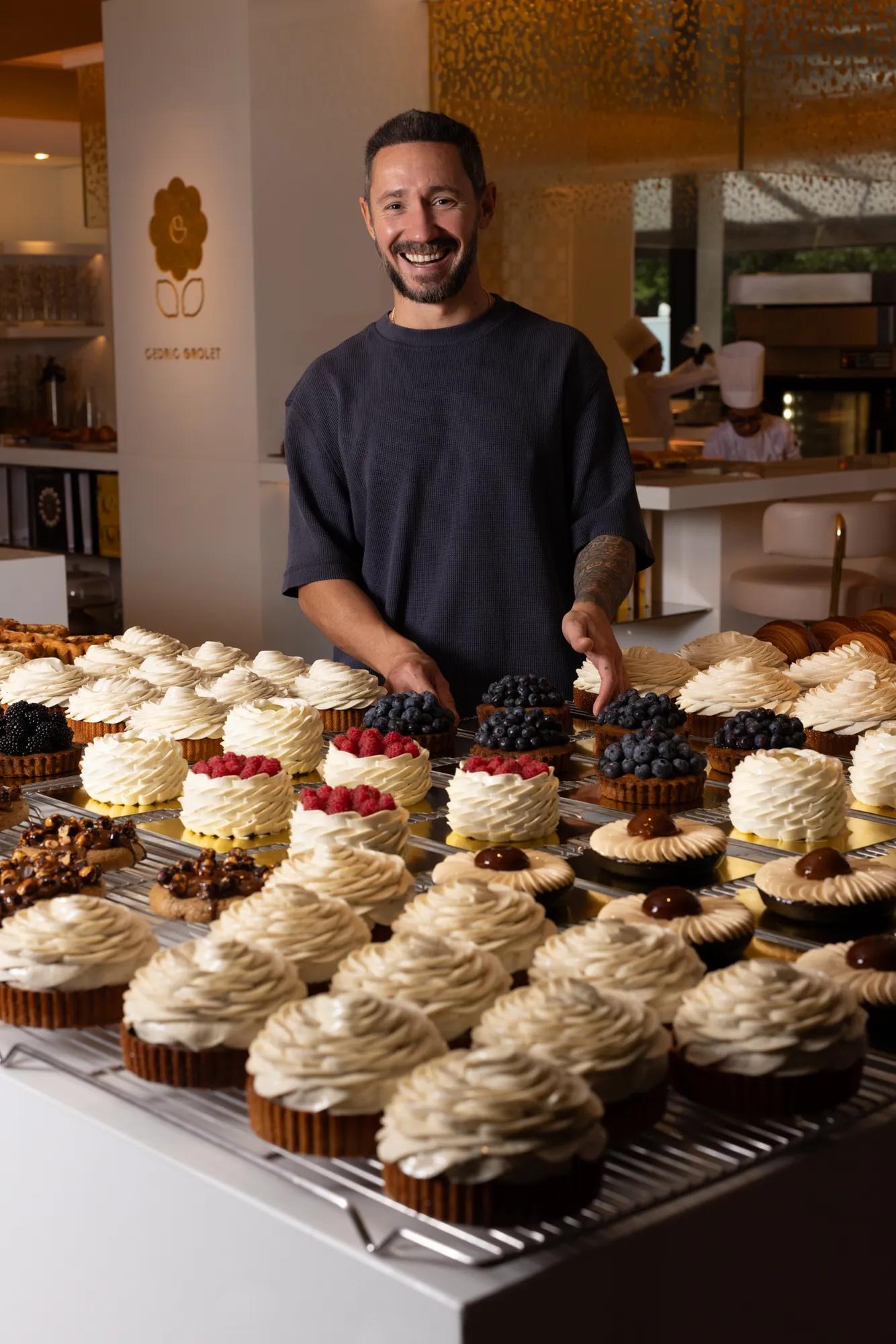 Pastry chef standing behind a display of cream-topped cakes and fruit pastries in a warm-lit bakery kitchen.
