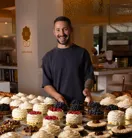 Pastry chef standing behind a display of cream-topped cakes and fruit pastries in a warm-lit bakery kitchen.
