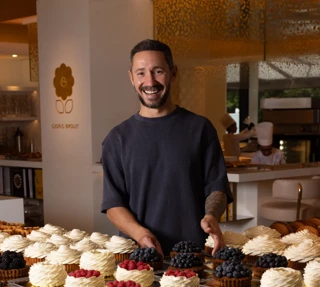 Pastry chef standing behind a display of cream-topped cakes and fruit pastries in a warm-lit bakery kitchen.